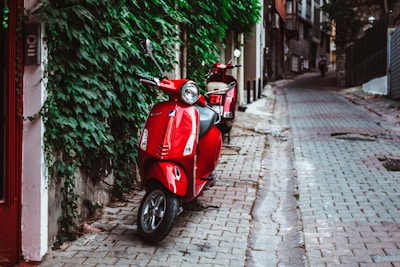 A red scooter is parked on a cobblestone street beside a wall covered in green ivy. Another red scooter is visible in the background along the narrow street bordered by buildings. The scene is quiet, with a solitary figure walking in the distance.