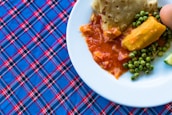 A plate of food resting on a plaid patterned tablecloth. The plate contains a dish with peas, an orange root vegetable, and pieces of fried flatbread partially soaked in a red sauce.