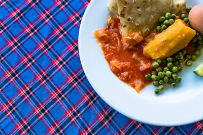 A plate of food resting on a plaid patterned tablecloth. The plate contains a dish with peas, an orange root vegetable, and pieces of fried flatbread partially soaked in a red sauce.