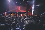 A large crowd of people enjoying a live concert in an indoor venue. The stage is illuminated with vibrant lights, and performers are visible on stage, with a screen displaying 'TEDASHII' behind them. The audience is engaged, with hands raised towards the stage.