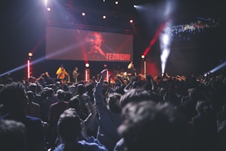 A large crowd of people enjoying a live concert in an indoor venue. The stage is illuminated with vibrant lights, and performers are visible on stage, with a screen displaying 'TEDASHII' behind them. The audience is engaged, with hands raised towards the stage.