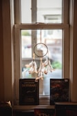A dreamcatcher with light brown feathers and a woven web hangs in front of a window, with natural light illuminating it from behind. On the windowsill below, there are two books. One book features the title 'Dothraki, Game of Thrones', and the other is titled 'Swordmaster'.