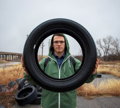 A person in a green jacket stands outdoors, holding a large tire with both hands. The landscape is slightly overcast with dry grass and a few trees in the background. A road and other tires are visible behind the person.
