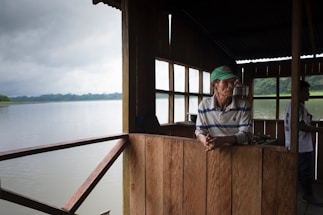 An elderly man wearing a green cap and striped shirt is leaning against a wooden partition inside a structure with open windows overlooking a serene body of water. The interior is dimly lit, and the scenery outside includes a cloudy sky and distant forested hills.