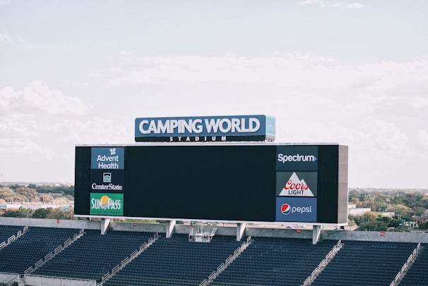A large digital scoreboard displays various advertisements at Camping World Stadium. The scoreboard is positioned in front of rows of empty stadium seats, and the background features a clear, slightly cloudy sky and some distant scenery including trees and buildings.