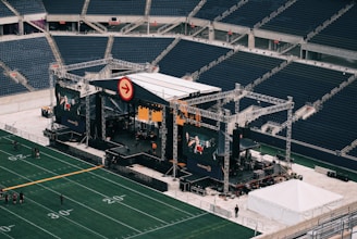 A large outdoor stage is set up inside a stadium, equipped with tall scaffolding, lights, and multiple large display screens. The seats in the stadium are mostly empty. The field is marked with numbers and lines typical of a sports venue. A few people are visible near the stage and on the field.