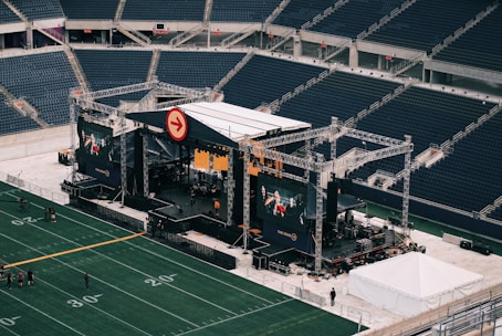 A large outdoor stage is set up inside a stadium, equipped with tall scaffolding, lights, and multiple large display screens. The seats in the stadium are mostly empty. The field is marked with numbers and lines typical of a sports venue. A few people are visible near the stage and on the field.