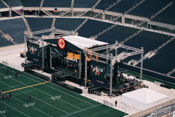 A large outdoor stage is set up inside a stadium, equipped with tall scaffolding, lights, and multiple large display screens. The seats in the stadium are mostly empty. The field is marked with numbers and lines typical of a sports venue. A few people are visible near the stage and on the field.