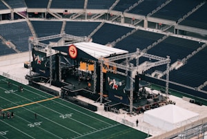 A large outdoor stage is set up inside a stadium, equipped with tall scaffolding, lights, and multiple large display screens. The seats in the stadium are mostly empty. The field is marked with numbers and lines typical of a sports venue. A few people are visible near the stage and on the field.