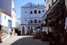 A vibrant street scene features several blue-painted buildings with ornate windows and doors, reminiscent of Moroccan architecture. The street is lined with various stalls displaying colorful textiles, leather goods, and artisanal crafts. The sky is clear and blue, adding to the bright and lively atmosphere of the setting.