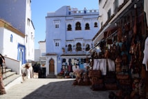 A vibrant street scene features several blue-painted buildings with ornate windows and doors, reminiscent of Moroccan architecture. The street is lined with various stalls displaying colorful textiles, leather goods, and artisanal crafts. The sky is clear and blue, adding to the bright and lively atmosphere of the setting.