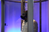 A person wearing a virtual reality headset is inside a glass enclosure. The scene is illuminated with a blue light, creating a futuristic ambience. The person is wearing a light-colored puffy jacket and stands facing sideways.
