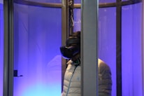 A person wearing a virtual reality headset is inside a glass enclosure. The scene is illuminated with a blue light, creating a futuristic ambience. The person is wearing a light-colored puffy jacket and stands facing sideways.