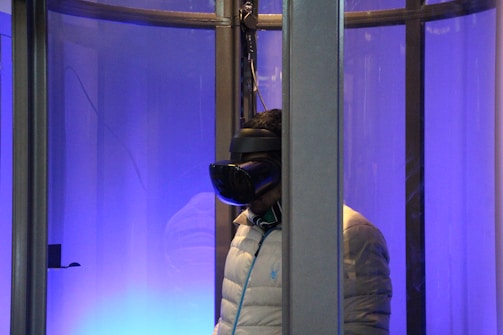 A person wearing a virtual reality headset is inside a glass enclosure. The scene is illuminated with a blue light, creating a futuristic ambience. The person is wearing a light-colored puffy jacket and stands facing sideways.