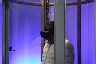 A person wearing a virtual reality headset is inside a glass enclosure. The scene is illuminated with a blue light, creating a futuristic ambience. The person is wearing a light-colored puffy jacket and stands facing sideways.