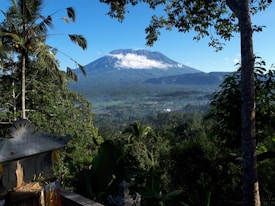 A lush, green landscape with dense tropical foliage in the foreground and a towering volcano in the background shrouded in wisps of white clouds. Blue skies and vibrant greenery enhance the natural beauty of the scene. A traditional stone structure with ornate carvings and small flags is visible partially in the bottom left corner.