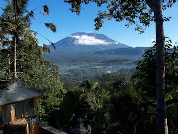 A lush, green landscape with dense tropical foliage in the foreground and a towering volcano in the background shrouded in wisps of white clouds. Blue skies and vibrant greenery enhance the natural beauty of the scene. A traditional stone structure with ornate carvings and small flags is visible partially in the bottom left corner.