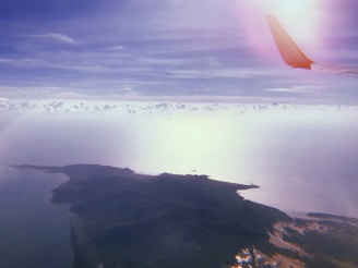 An aerial view of a landscape with expansive cloud cover and visible landmasses. The image is framed by an airplane wing on the top right corner, indicating it is taken from a flying aircraft. The atmosphere appears serene with a mix of light and shadows playing on the water and land below.