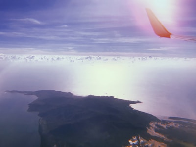 An aerial view of a landscape with expansive cloud cover and visible landmasses. The image is framed by an airplane wing on the top right corner, indicating it is taken from a flying aircraft. The atmosphere appears serene with a mix of light and shadows playing on the water and land below.