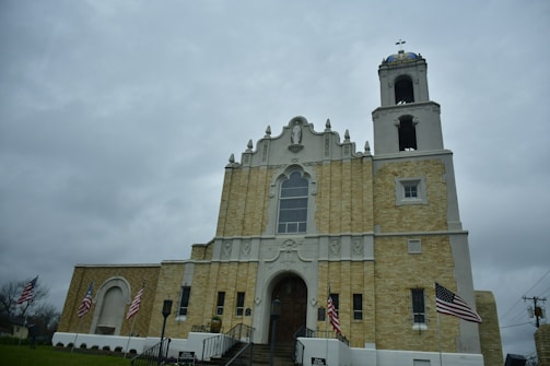A large, historic church with a beige brick facade and ornate architectural details stands prominently. The building features a tall bell tower with a dome and cross on top. Several American flags are displayed around the entrance, which is accessed by a set of stairs with railings. The sky above is overcast.
