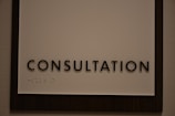 A sign featuring the word 'CONSULTATION' in bold, black capital letters. Beneath the text, there is Braille marked on a white background, surrounded by a dark wooden frame.