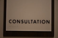 A sign featuring the word 'CONSULTATION' in bold, black capital letters. Beneath the text, there is Braille marked on a white background, surrounded by a dark wooden frame.