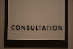 A sign featuring the word 'CONSULTATION' in bold, black capital letters. Beneath the text, there is Braille marked on a white background, surrounded by a dark wooden frame.