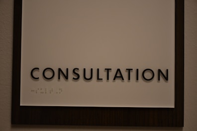 A sign featuring the word 'CONSULTATION' in bold, black capital letters. Beneath the text, there is Braille marked on a white background, surrounded by a dark wooden frame.