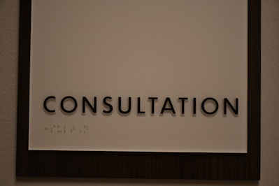 A sign featuring the word 'CONSULTATION' in bold, black capital letters. Beneath the text, there is Braille marked on a white background, surrounded by a dark wooden frame.
