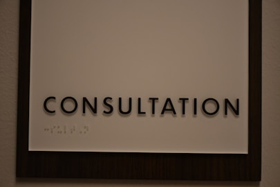 A sign featuring the word 'CONSULTATION' in bold, black capital letters. Beneath the text, there is Braille marked on a white background, surrounded by a dark wooden frame.