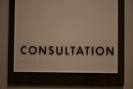 A sign featuring the word 'CONSULTATION' in bold, black capital letters. Beneath the text, there is Braille marked on a white background, surrounded by a dark wooden frame.