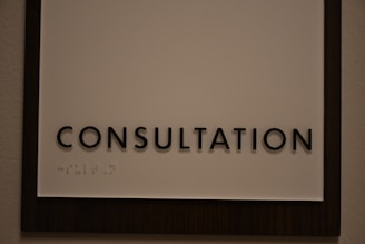 A sign featuring the word 'CONSULTATION' in bold, black capital letters. Beneath the text, there is Braille marked on a white background, surrounded by a dark wooden frame.