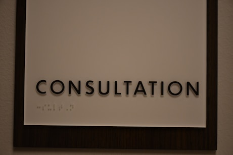 A sign featuring the word 'CONSULTATION' in bold, black capital letters. Beneath the text, there is Braille marked on a white background, surrounded by a dark wooden frame.