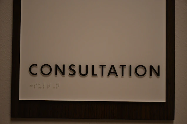A sign featuring the word 'CONSULTATION' in bold, black capital letters. Beneath the text, there is Braille marked on a white background, surrounded by a dark wooden frame.
