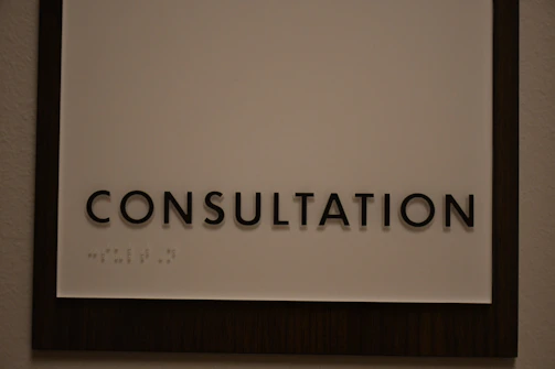 A sign featuring the word 'CONSULTATION' in bold, black capital letters. Beneath the text, there is Braille marked on a white background, surrounded by a dark wooden frame.