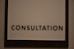 A sign featuring the word 'CONSULTATION' in bold, black capital letters. Beneath the text, there is Braille marked on a white background, surrounded by a dark wooden frame.
