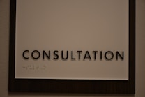 A sign featuring the word 'CONSULTATION' in bold, black capital letters. Beneath the text, there is Braille marked on a white background, surrounded by a dark wooden frame.