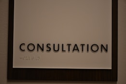A sign featuring the word 'CONSULTATION' in bold, black capital letters. Beneath the text, there is Braille marked on a white background, surrounded by a dark wooden frame.