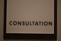 A sign featuring the word 'CONSULTATION' in bold, black capital letters. Beneath the text, there is Braille marked on a white background, surrounded by a dark wooden frame.
