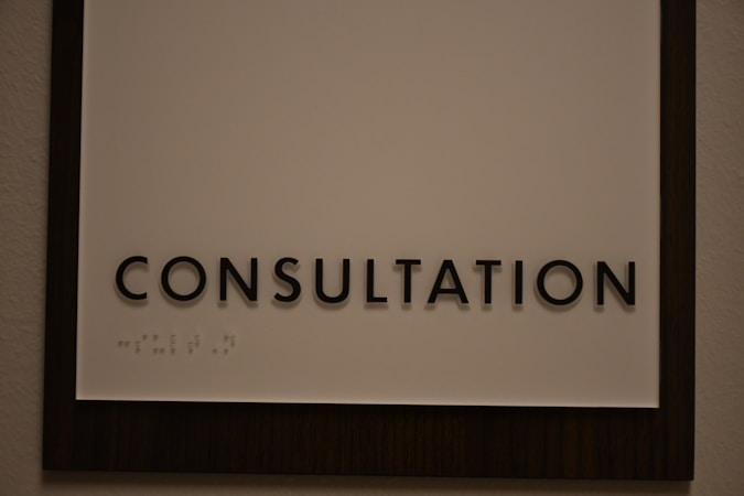 A sign featuring the word 'CONSULTATION' in bold, black capital letters. Beneath the text, there is Braille marked on a white background, surrounded by a dark wooden frame.