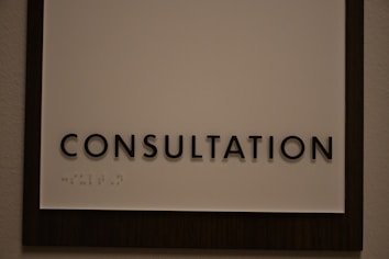 A sign featuring the word 'CONSULTATION' in bold, black capital letters. Beneath the text, there is Braille marked on a white background, surrounded by a dark wooden frame.