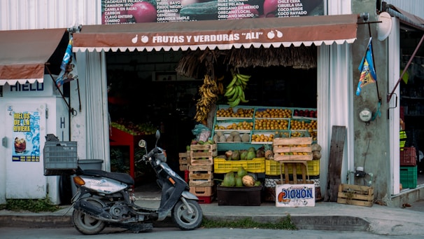A small market stall with a sign reading 'Frutas y Verduras Ali' that sells various fruits and vegetables. There are bananas, coconuts, and other fruits arranged in yellow crates outside the stall. A scooter is parked in front of the stall. The stall is partially enclosed with a white and brown awning, and there are signs and posters on the walls around it.