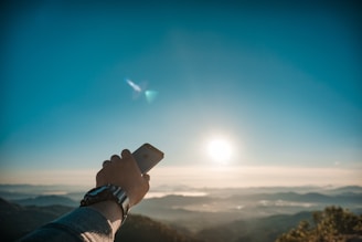 A hand holding a smartphone outdoors with a scenic mountain background.