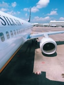 A passenger airplane, identified by the 'SilkAir' logo, is parked on the tarmac at an airport. The view highlights the aircraft's exterior, including its windows and a jet engine. The background features airport facilities and clear blue skies with scattered clouds.