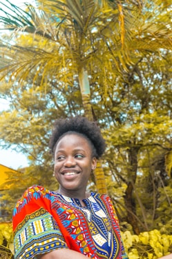 A joyful woman wearing colorful tropical print clothing, standing on a sunny beach with palm trees.