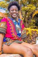 A person is sitting outdoors, wearing a vibrant, colorful traditional African dashiki. The fabric features intricate patterns in red, blue, and yellow. The person is smiling, exuding happiness and warmth. The background includes lush green foliage and a palm tree, suggesting a tropical environment.