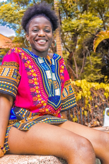 A person is sitting outdoors, wearing a vibrant, colorful traditional African dashiki. The fabric features intricate patterns in red, blue, and yellow. The person is smiling, exuding happiness and warmth. The background includes lush green foliage and a palm tree, suggesting a tropical environment.