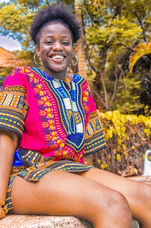 A person is sitting outdoors, wearing a vibrant, colorful traditional African dashiki. The fabric features intricate patterns in red, blue, and yellow. The person is smiling, exuding happiness and warmth. The background includes lush green foliage and a palm tree, suggesting a tropical environment.