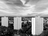 Three tall residential buildings are prominently situated in an urban area, surrounded by trees and a variety of smaller buildings and structures. The skyline is expansive, with a cloudy sky above casting varied shadows and highlights across the scene.