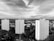 Three tall residential buildings are prominently situated in an urban area, surrounded by trees and a variety of smaller buildings and structures. The skyline is expansive, with a cloudy sky above casting varied shadows and highlights across the scene.
